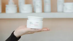 Hand holding a white jar with shelves of similar jars in the background