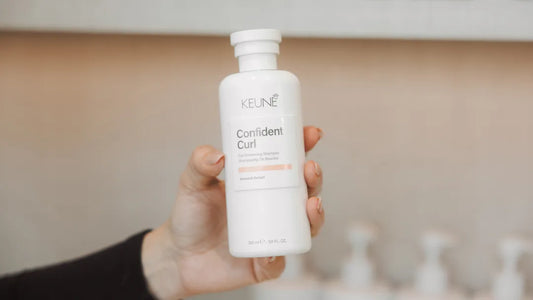 Hand holding a Keune Confident Curl product bottle against a blurred bathroom background