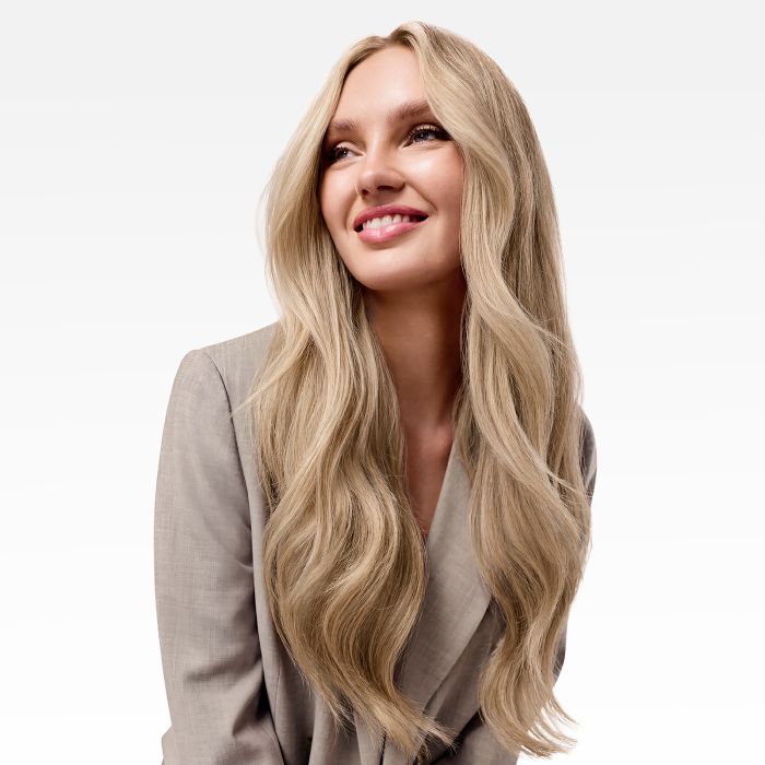 A woman with long, wavy blonde hair wearing a beige blazer, shown from the chest up against a clean white background.