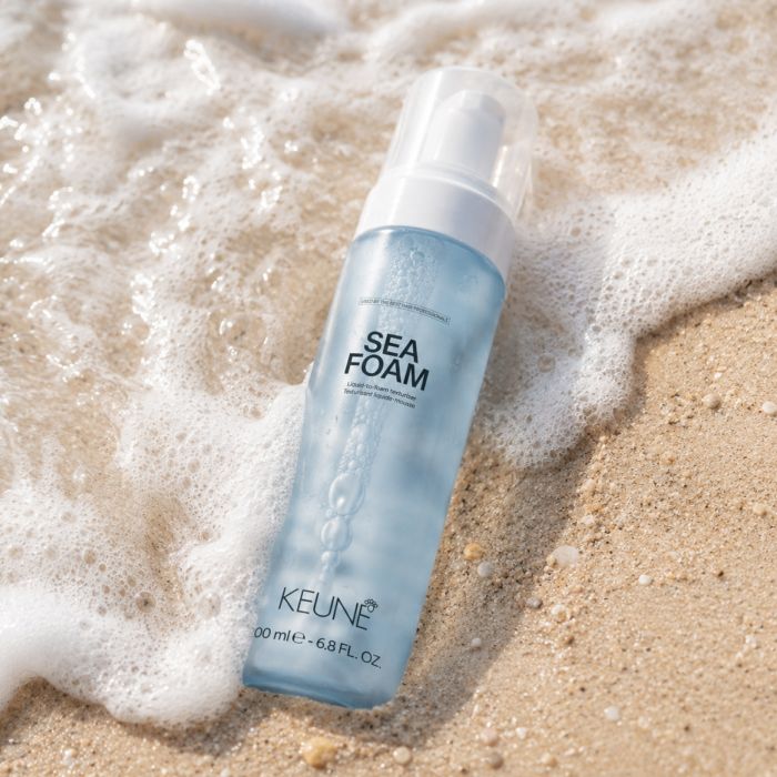 A bottle of KEUNE Sea Foam hair foam lies on sandy beach ground with foamy ocean water around it. The bottle is translucent light blue with a white pump and clear cap.