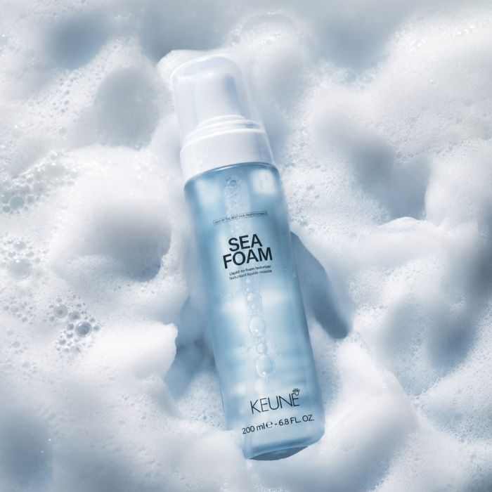 A bottle of KEUNE Sea Foam foam with a white pump, set against a background of white and light blue lathered foam.