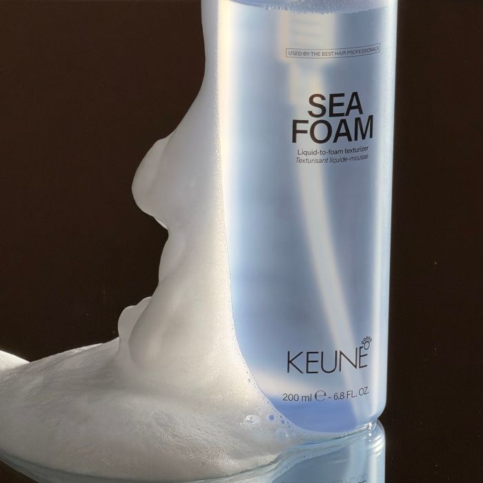 A bottle of KEUNE Sea Foam liquid-to-foam treatment with thick white foam dripping from the side, set against a dark brown background.