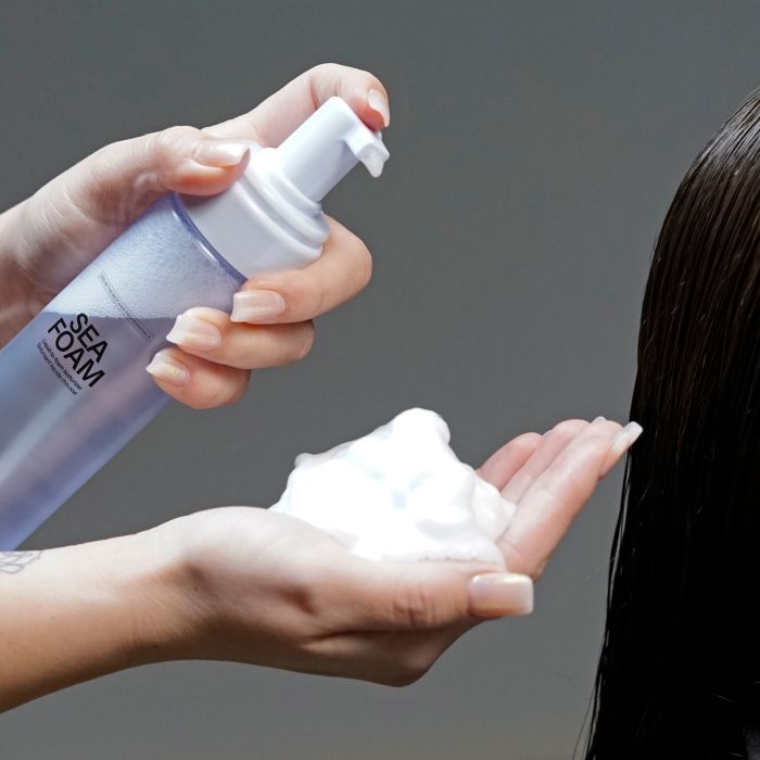 A close-up of a person applying Sea Foam  foam from a translucent bottle onto their hand, with dark hair visible on the right against a plain gray background.