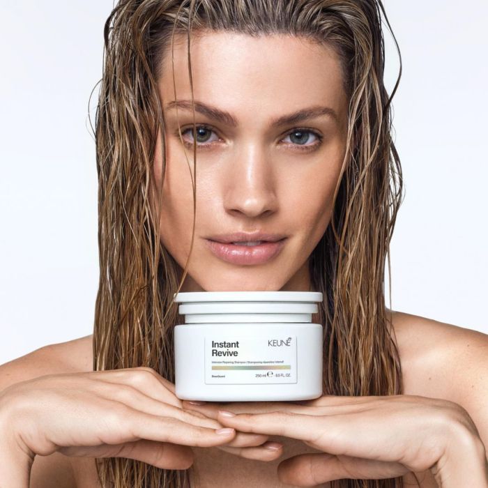 A wet woman holding a white jar of Keune Instant Revive hair repair mask against a clean white background.