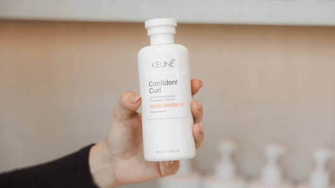 Hand holding a Keune Confident Curl product bottle against a blurred bathroom background
