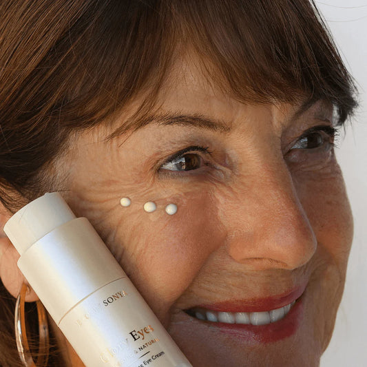 Woman holding a bottle of eye cream with visible droplets on her face.