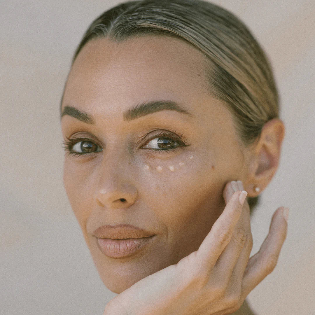 A close-up portrait of a woman with her hand resting on her cheek, showcasing a small, clear, round cosmetic shimmer or dot on her cheek. The background is a soft, neutral beige.