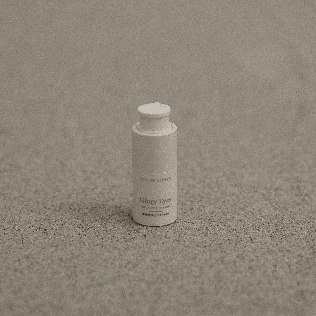 A small white cylindrical cosmetic bottle labeled 'ECO BY SONYA Glory Eyes' standing upright on a textured gray surface.
