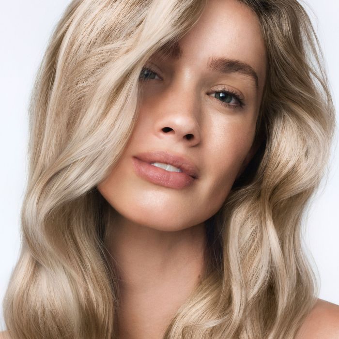 A close-up portrait of a woman with long, wavy blonde hair, showcasing a light golden blonde color with subtle warm brown undertones.