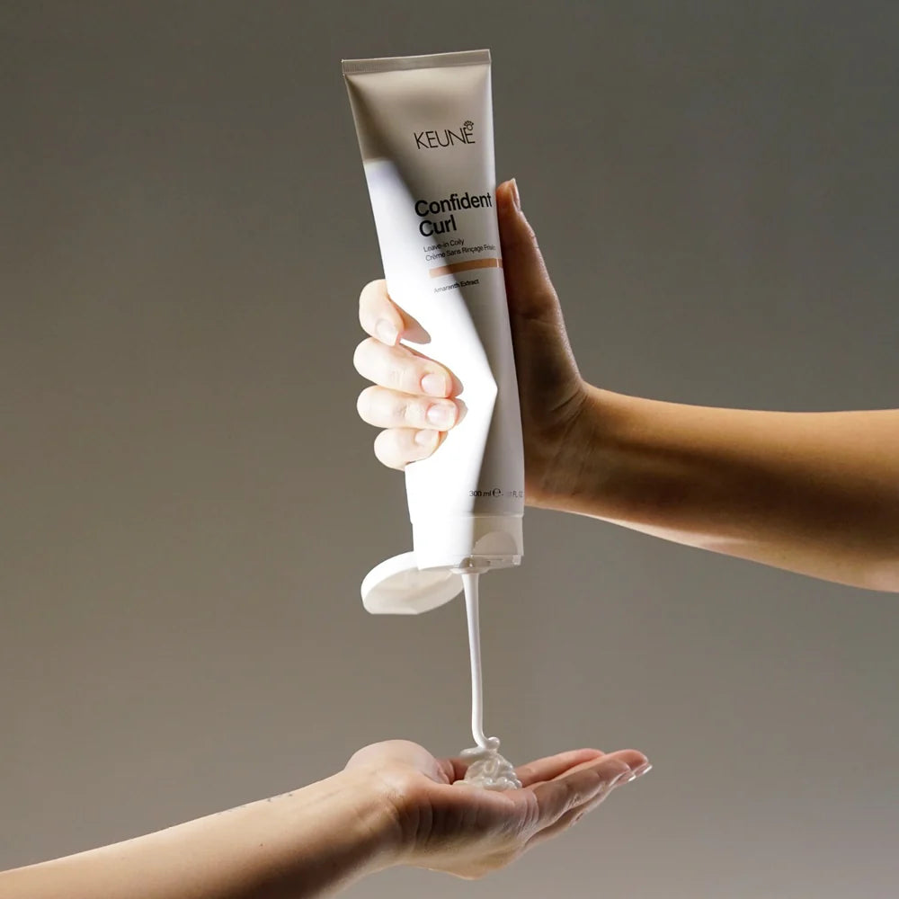 Hand holding a tube of Keune Confidenc Curl product with visible cream being squeezed out on a neutral background