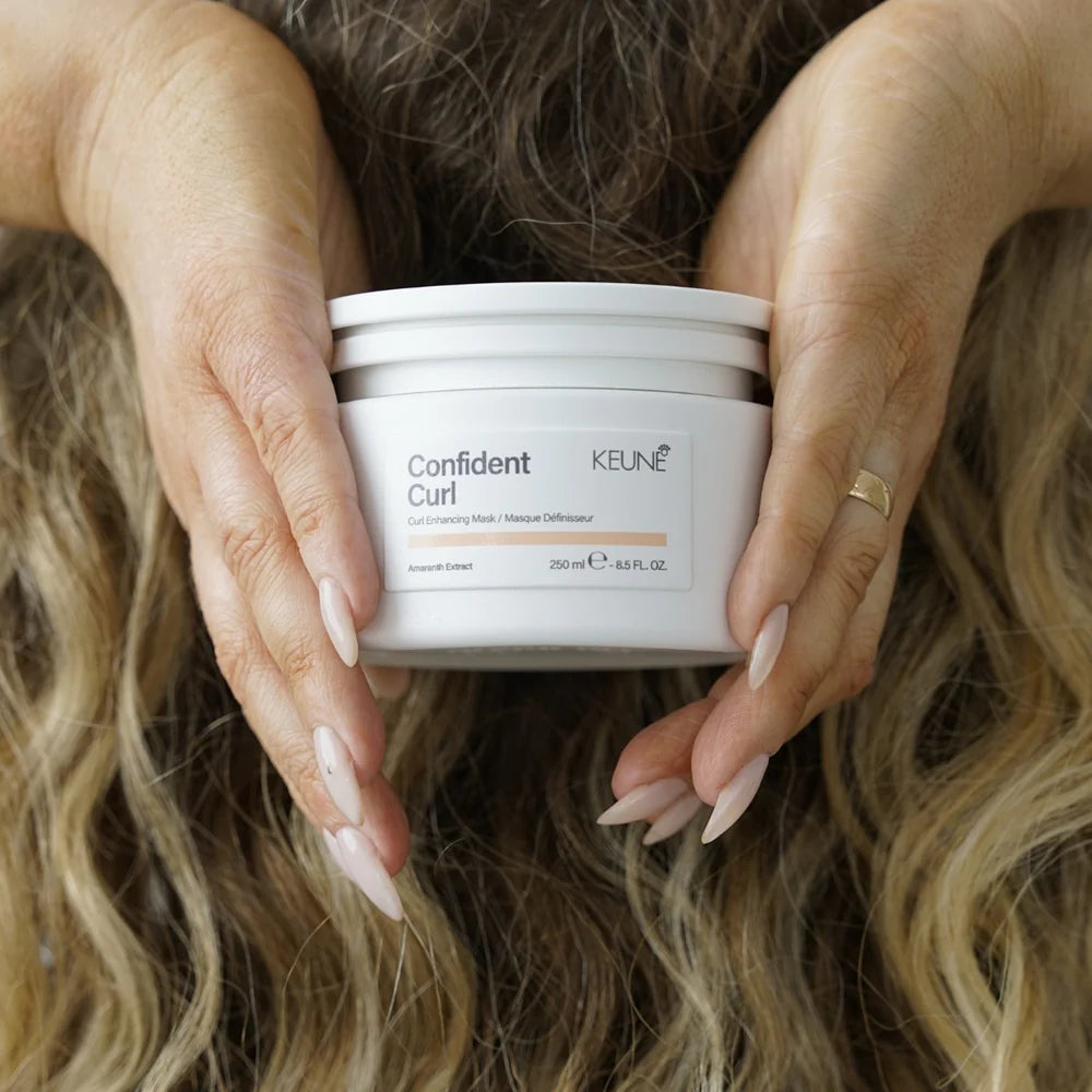 Person holding a jar of Keune Confident Curl hair product with wavy hair in the background