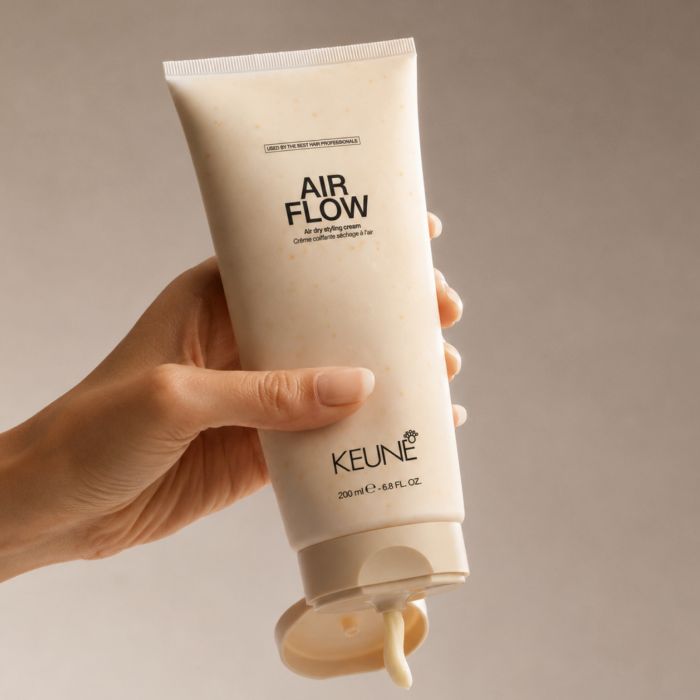A hand holding a squeeze tube of KEUNE Air Flow hair oil against a neutral beige background.