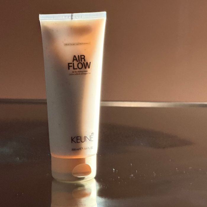 A 200ml squeeze tube of KEUNE Air Flow hair styling cream standing upright on a reflective dark surface against a warm brown background.