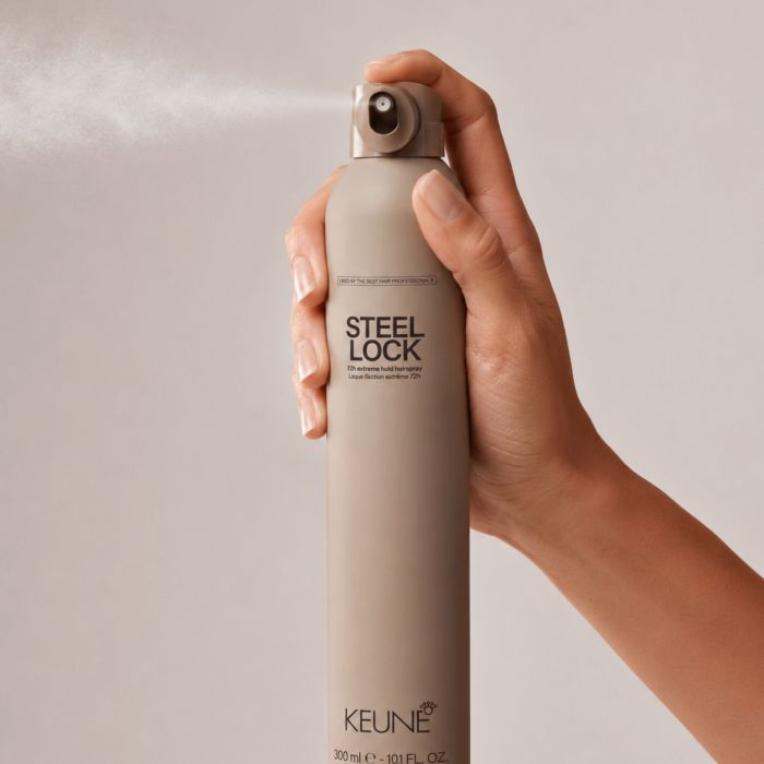 A hand holding a can of KEUNE Steel Lock hair styling spray, with the spray nozzle activated and mist visible against a plain light beige background.