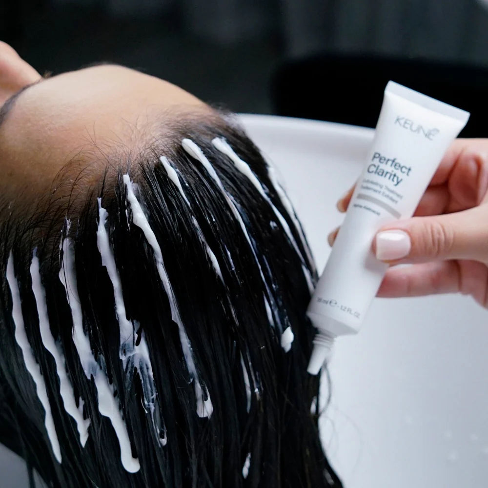 Person with black hair being washed, holding a tube of Keune Perfect Clarity product.