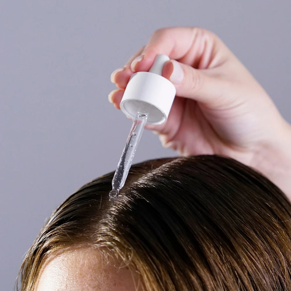 Person applying a clear liquid to their hair using a dropper bottle against a gray background